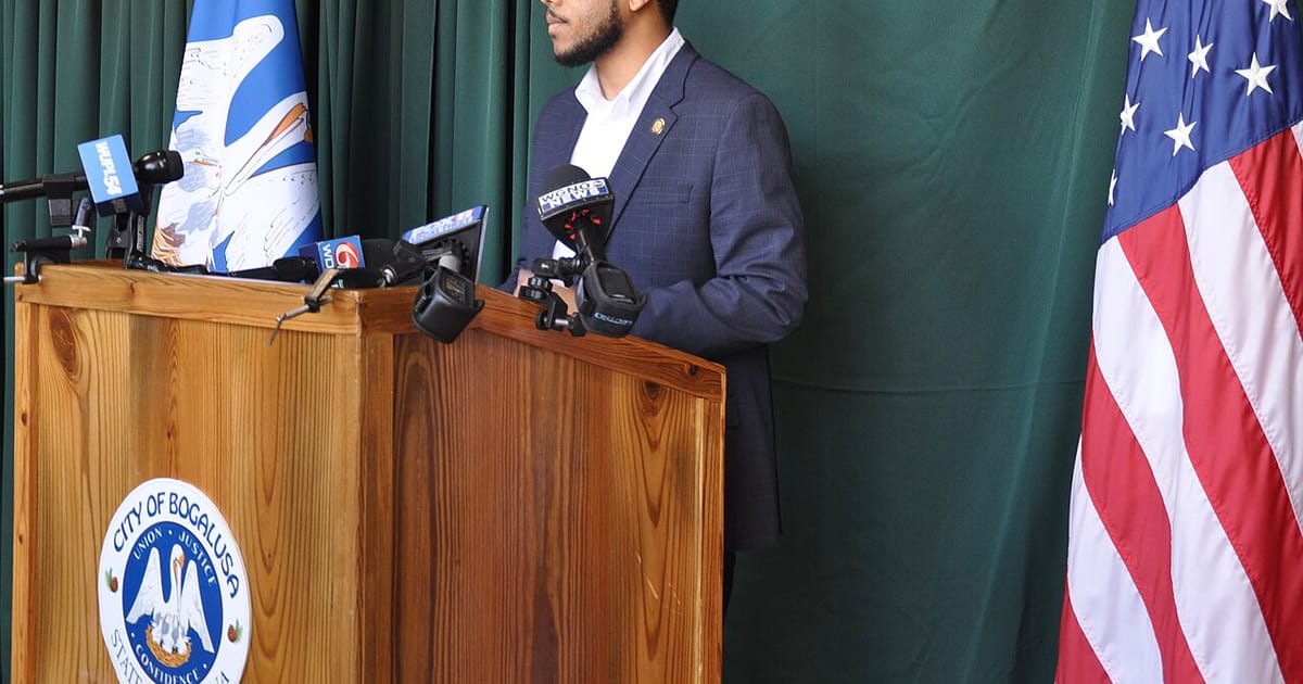 A speaker at a podium facing a cluster of television news microphones